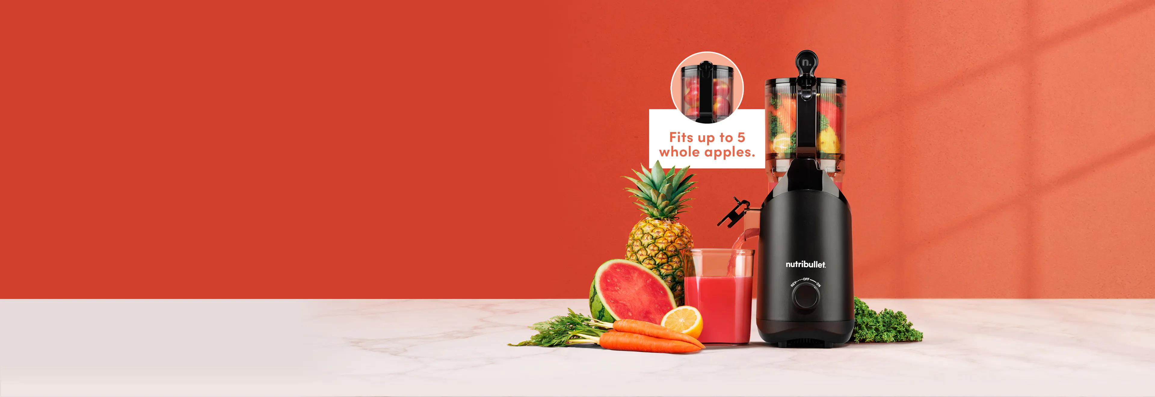 Black nutribullet Cold Press Juicer filled with carrots, kale, and lemon, pouring red juice into a glass next to watermelon, pineapple, and carrots on a marble counter.