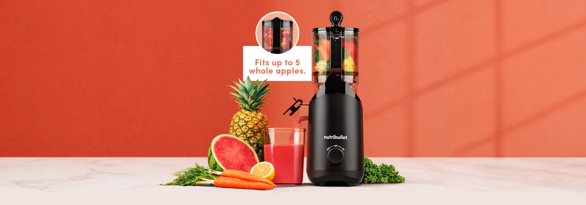 Black nutribullet Cold Press Juicer filled with carrots, kale, and lemon, pouring red juice into a glass next to watermelon, pineapple, and carrots on a marble counter.