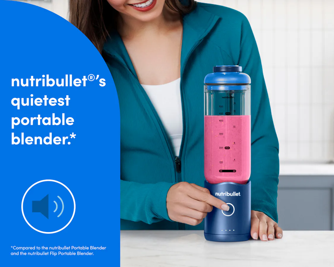 A woman in a teal zip-up athletic jacket presses the blend button on the bottom of the motor base to blend a bright pink smoothie.