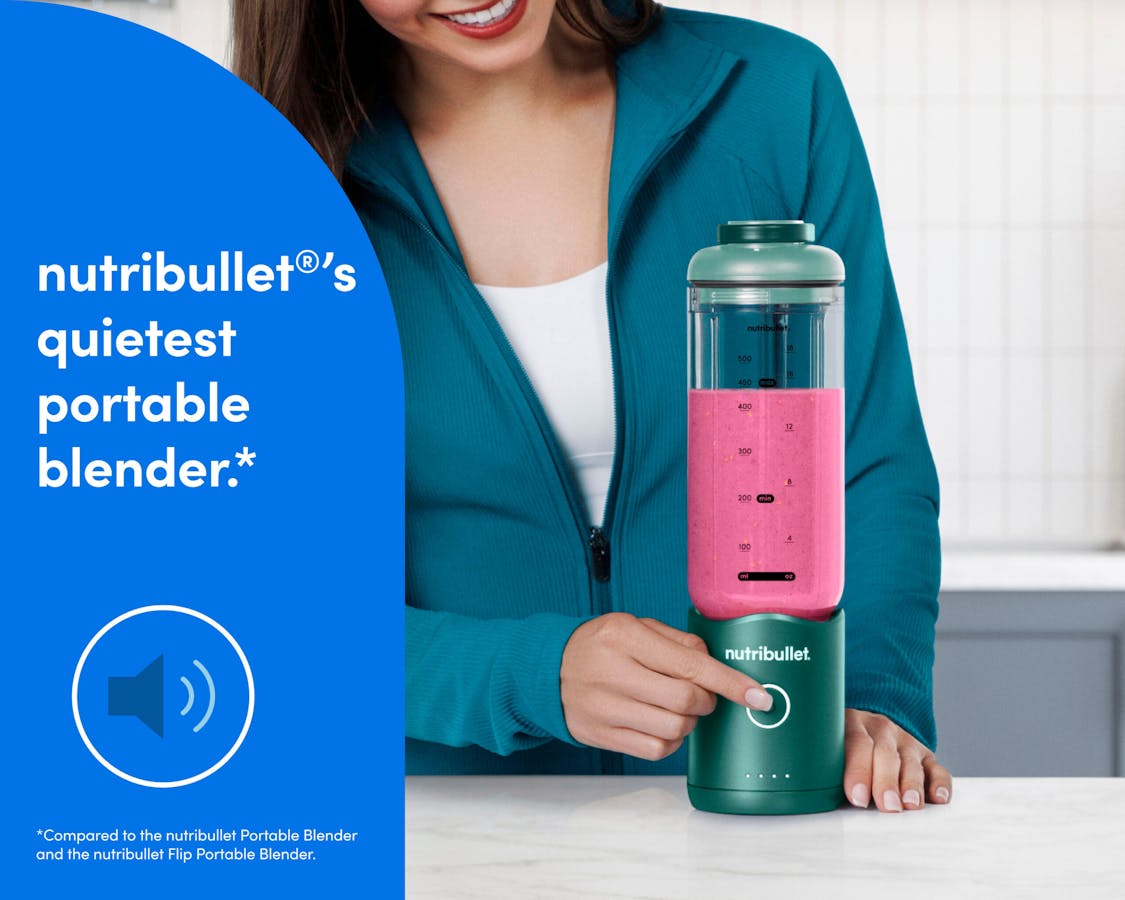 A woman in a teal zip-up athletic jacket presses the blend button on the bottom of the motor base to blend a bright pink smoothie.