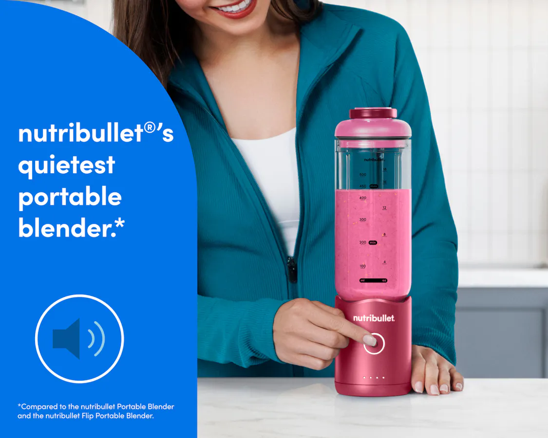 A woman in a teal zip-up athletic jacket presses the blend button on the bottom of the motor base to blend a bright pink smoothie.