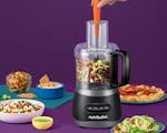 Product preview 3 of 8. Thumbnail of nutribullet® 7-Cup Food Processor with a carrot in its chute, filled with shredded vegetables surrounded by pizza, dips and salsa against a purple background