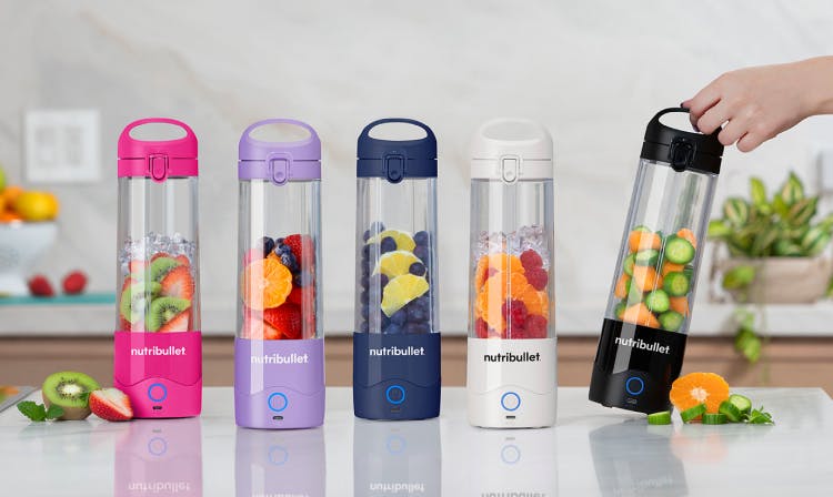 NutriBullet portable bottles in various colors (pink, purple, blue, white, and black) filled with fresh fruits and vegetables, placed on a kitchen countertop