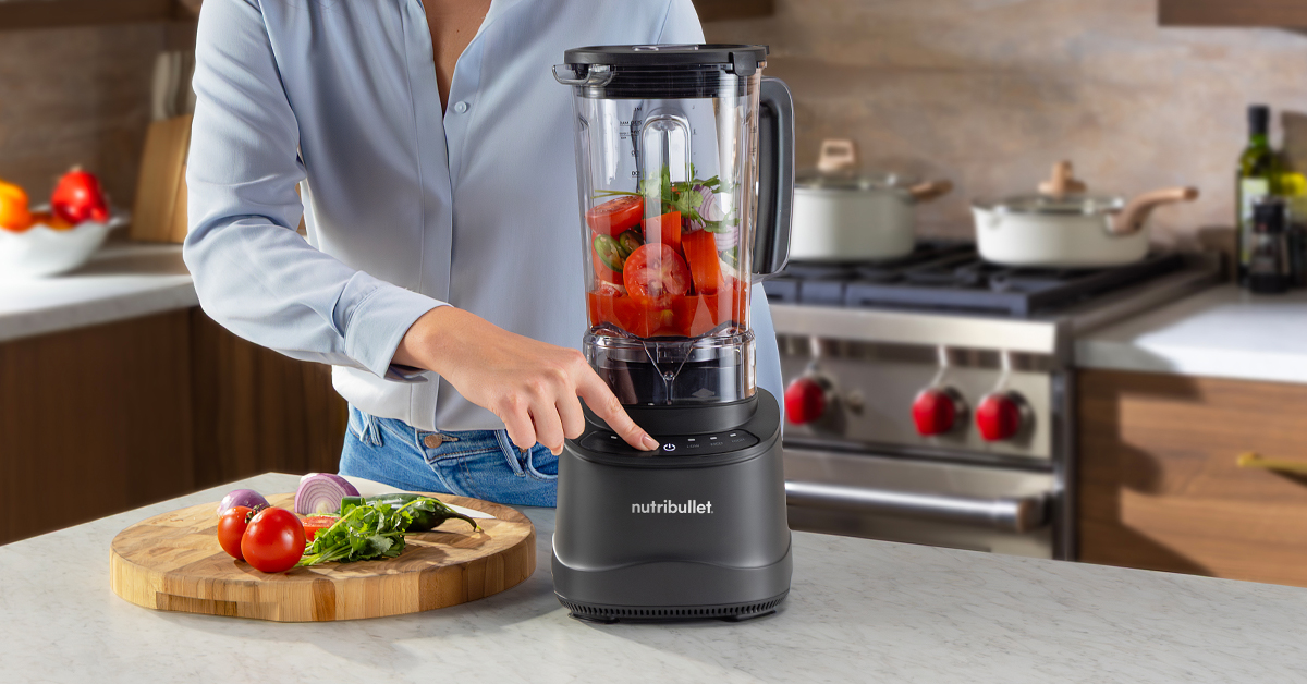 nutribullet PowerCore Blender on kitchen countertop filled with fresh tomatoes and herbs next to someone about to push "blend"