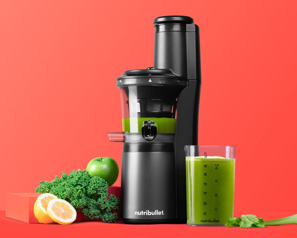 NEW black juicer filled with green juice next to cup of juice, fruits, veggies on orange background.