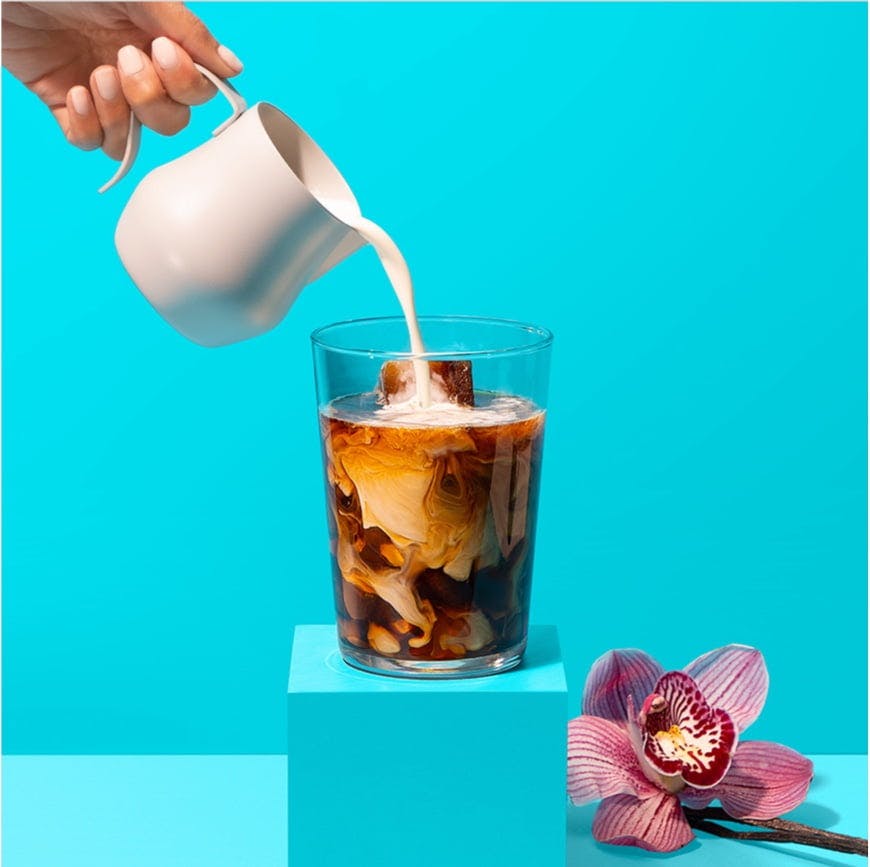 Hand pouring milk into glass of coffee on a pedestal with a flower on a blue background.