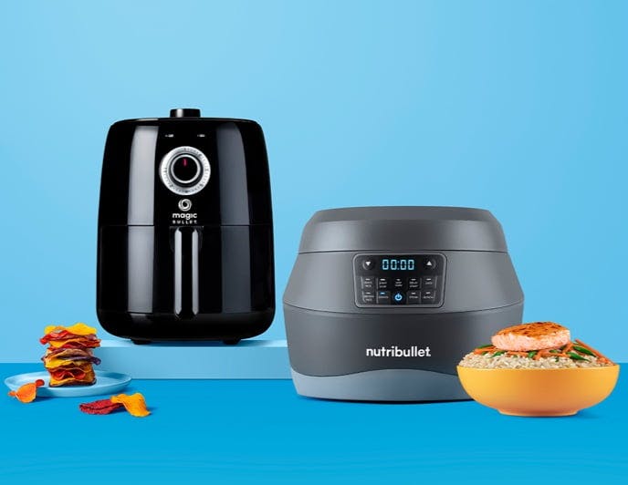 Black airfryer and gray EveryGrain Cooker on blue background, plates and bowls of food in foreground