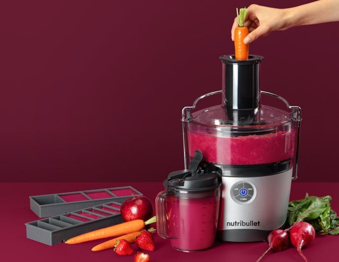 nutribullet juicer and accessories, cups, and ice trays with vegetables on maroon background.