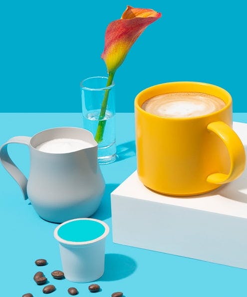 Yellow coffee mug, grey cup of milk, container of coffee grounds and flower vase on blue background.